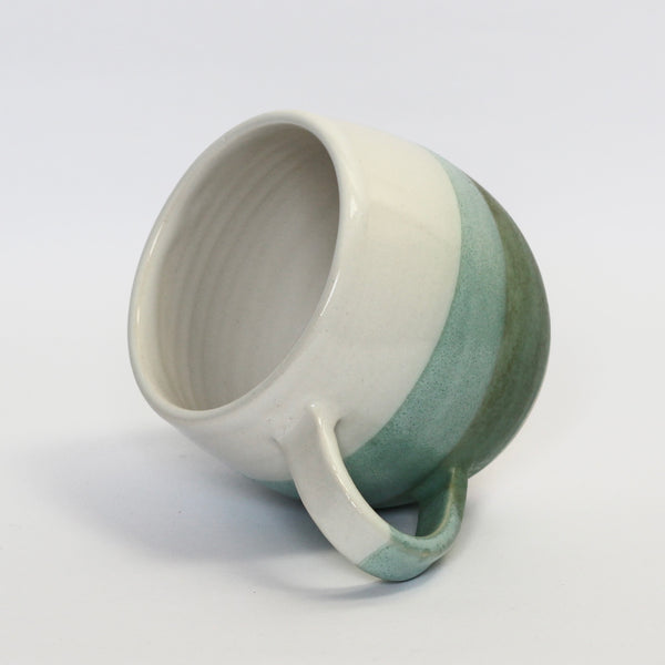 stoneware curved mug resting on it's handle, glazed in white and dark bluey, green. the overlap is a mid green