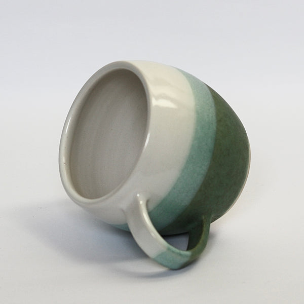 stoneware curved mug balanced on handle, glazed in white and dark bluey, green. the overlap is a mid green