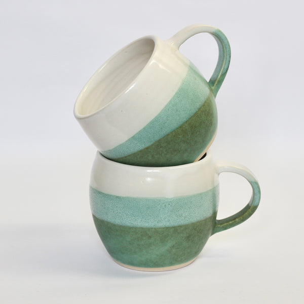 two stacked stoneware curved mugs, glazed in white and dark bluey, green. the overlap is a mid green